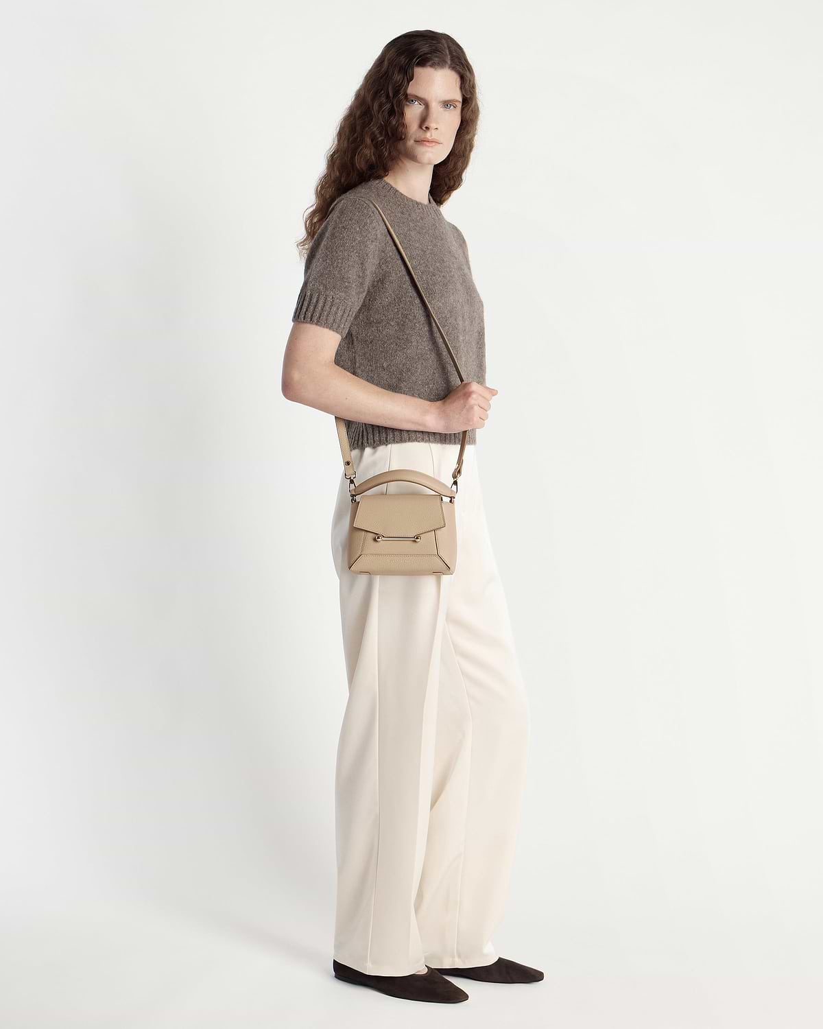 Model wearing a small sand-coloured structured top-handle crossbody bag with long strap over shoulder