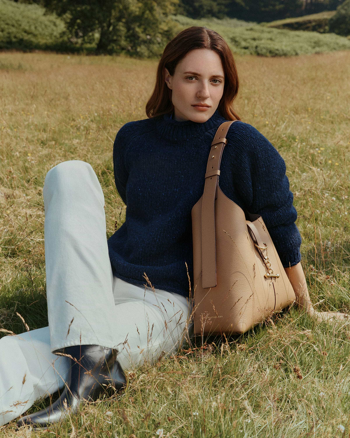 Woman sitting in field wearing navy jumper with caramel leather Kite Hobo Maxi bag.