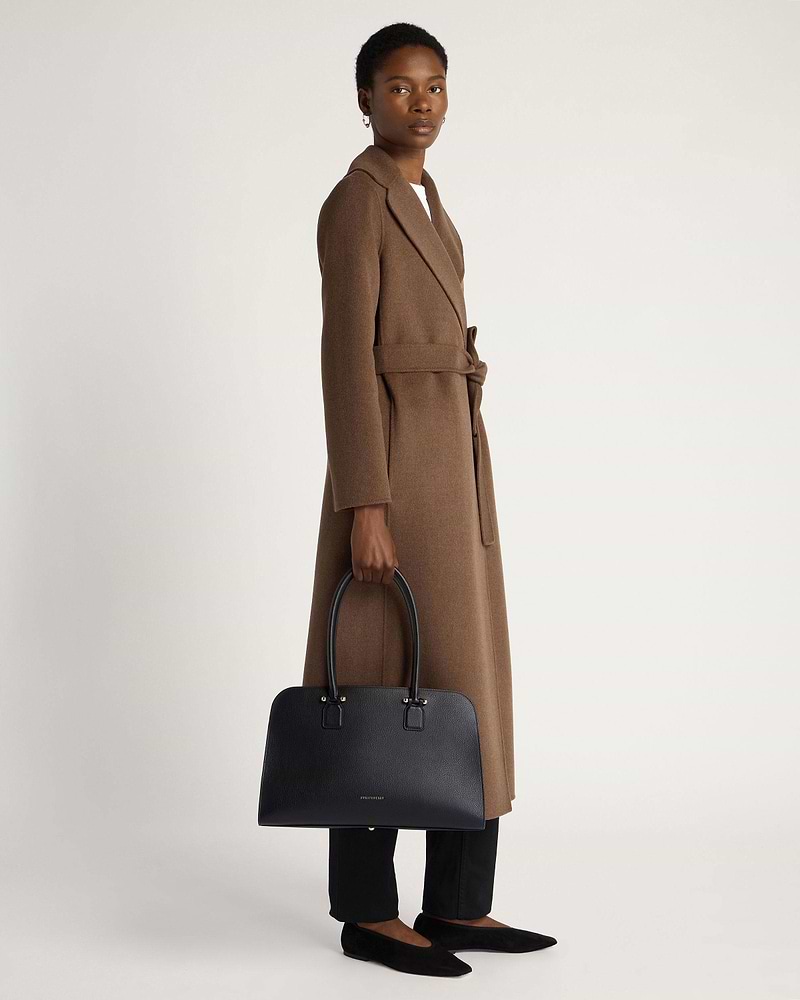 Woman holding black Georgia Maxi tote bag while wearing a brown belted coat.