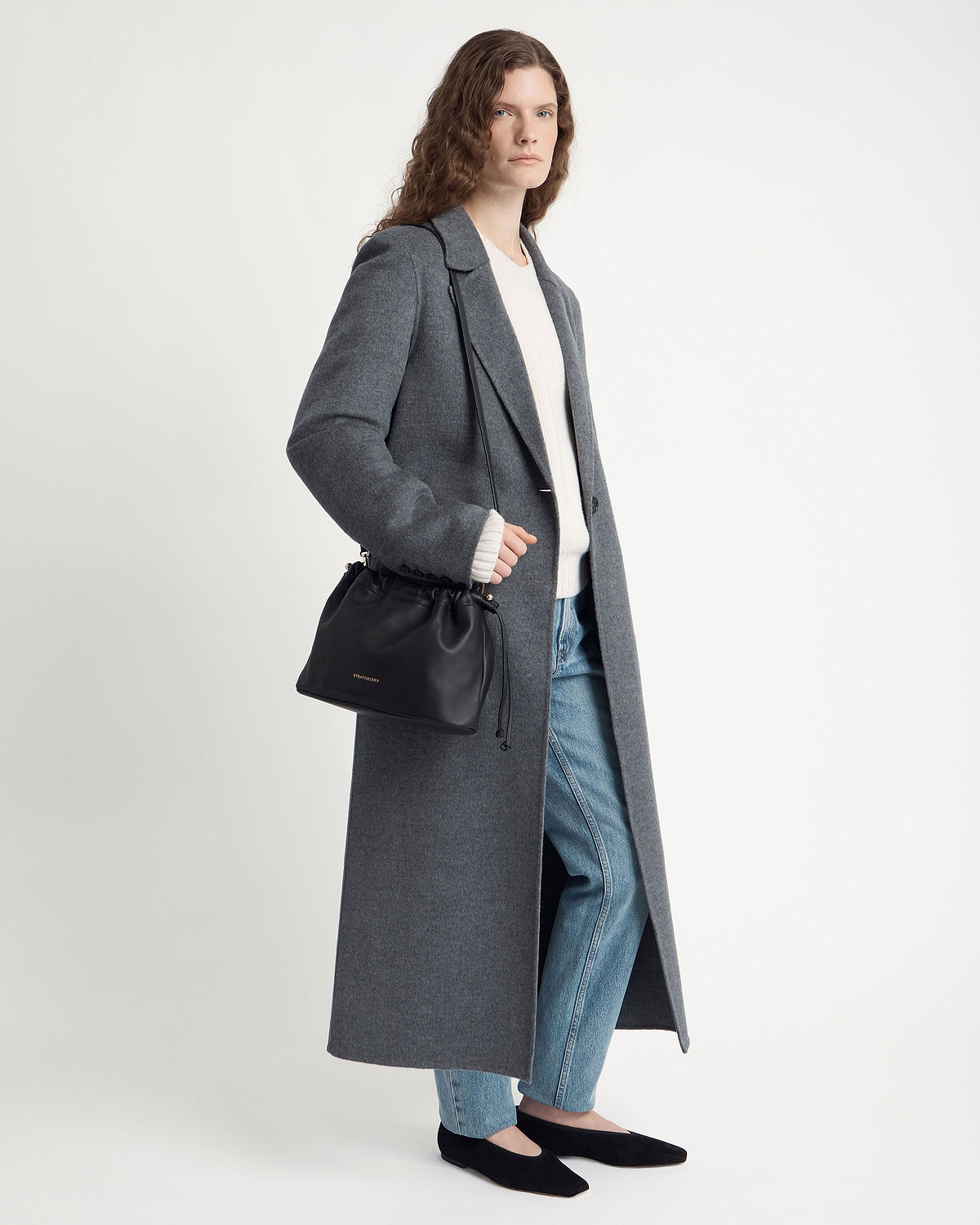 Model wearing a grey long coat and jeans carries a Strathberry black drawstring bag.
