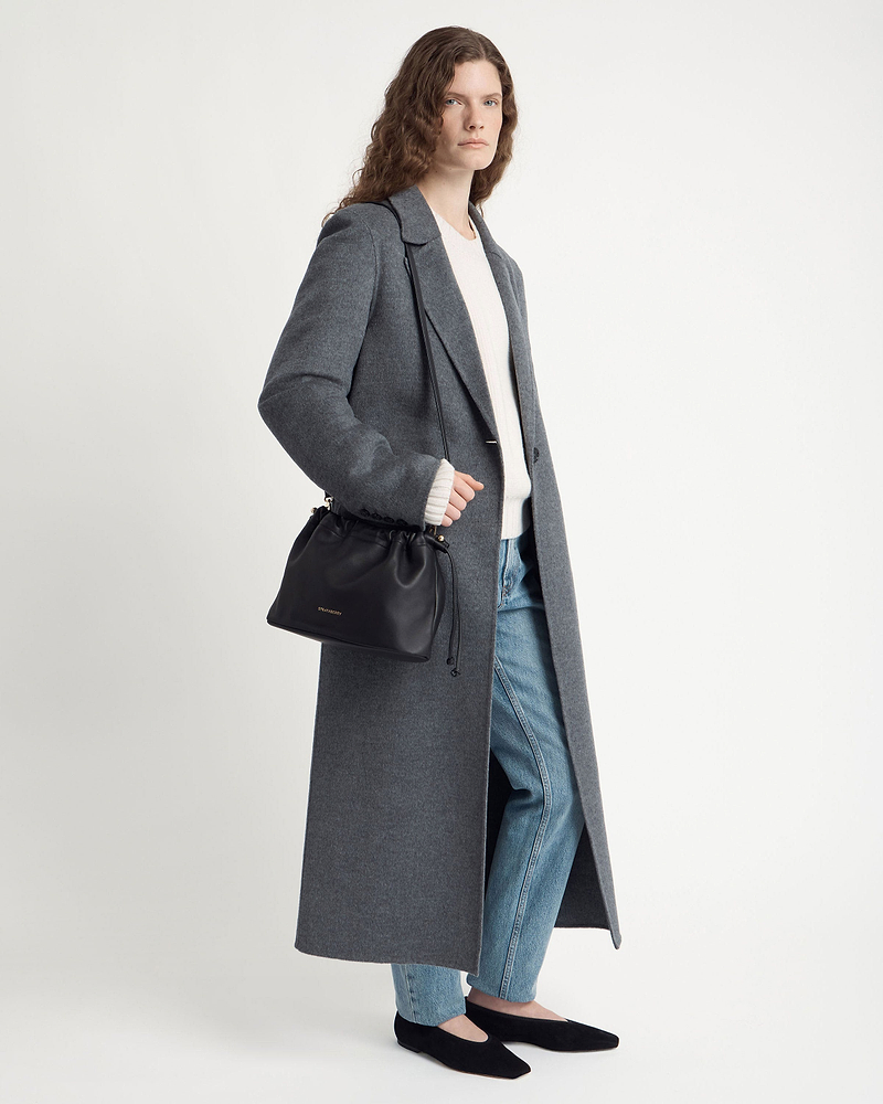Model wearing a grey long coat and jeans carries a Strathberry black drawstring bag.