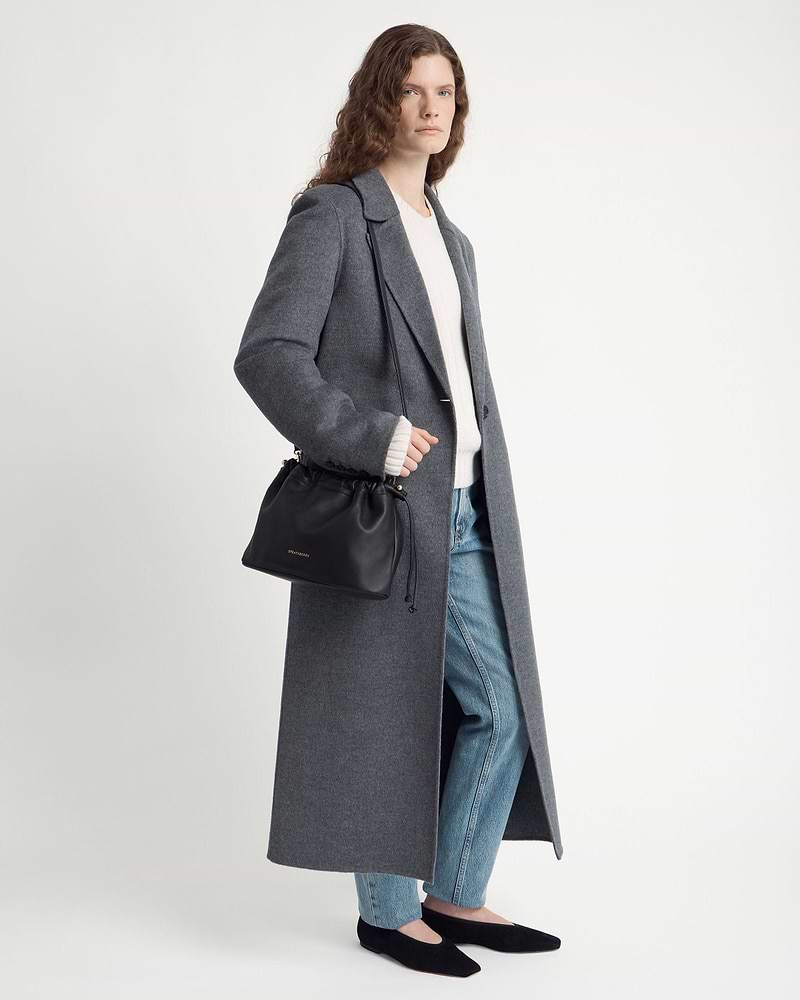 Model wearing a grey long coat and jeans carries a Strathberry black drawstring bag.