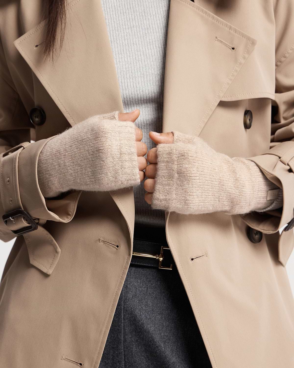 Hands in oat fingerless cashmere gloves holding beige trench coat lapels.