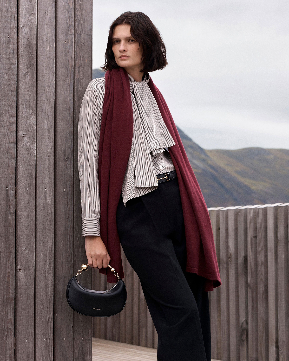 Model holding black Strathberry Crescent Moon Mini bag by gold handle, wearing burgundy scarf.