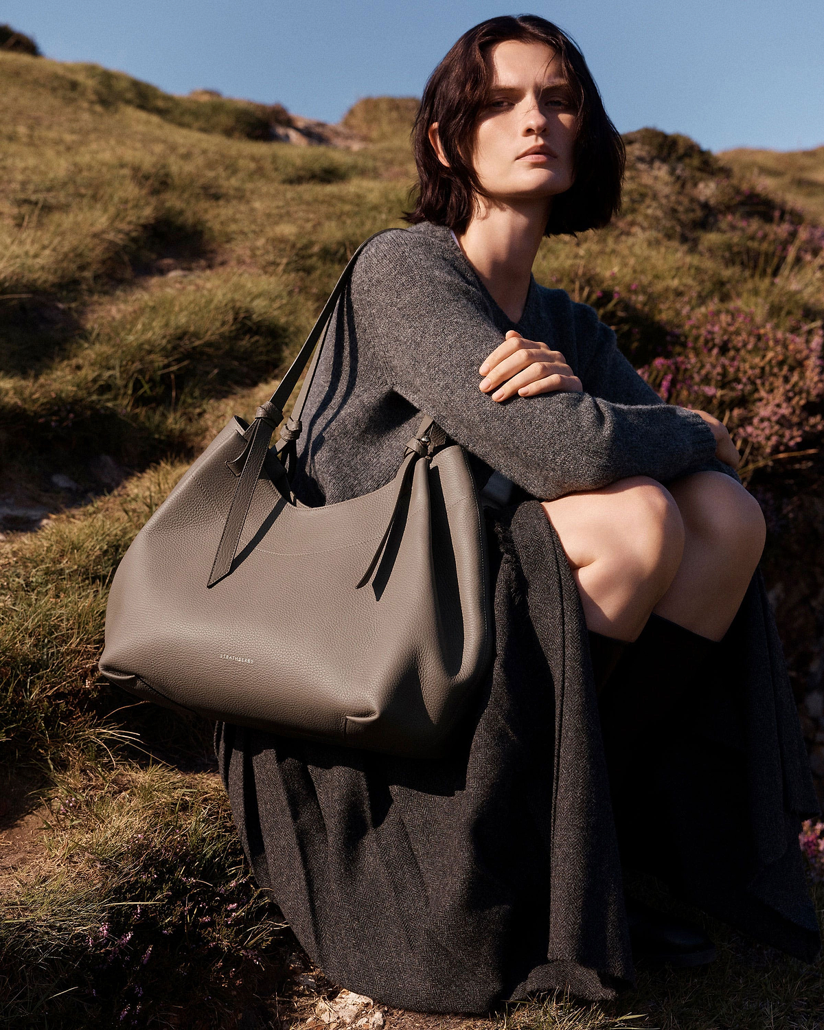 Model sitting outdoors holding taupe Strathberry Barra tote over shoulder.