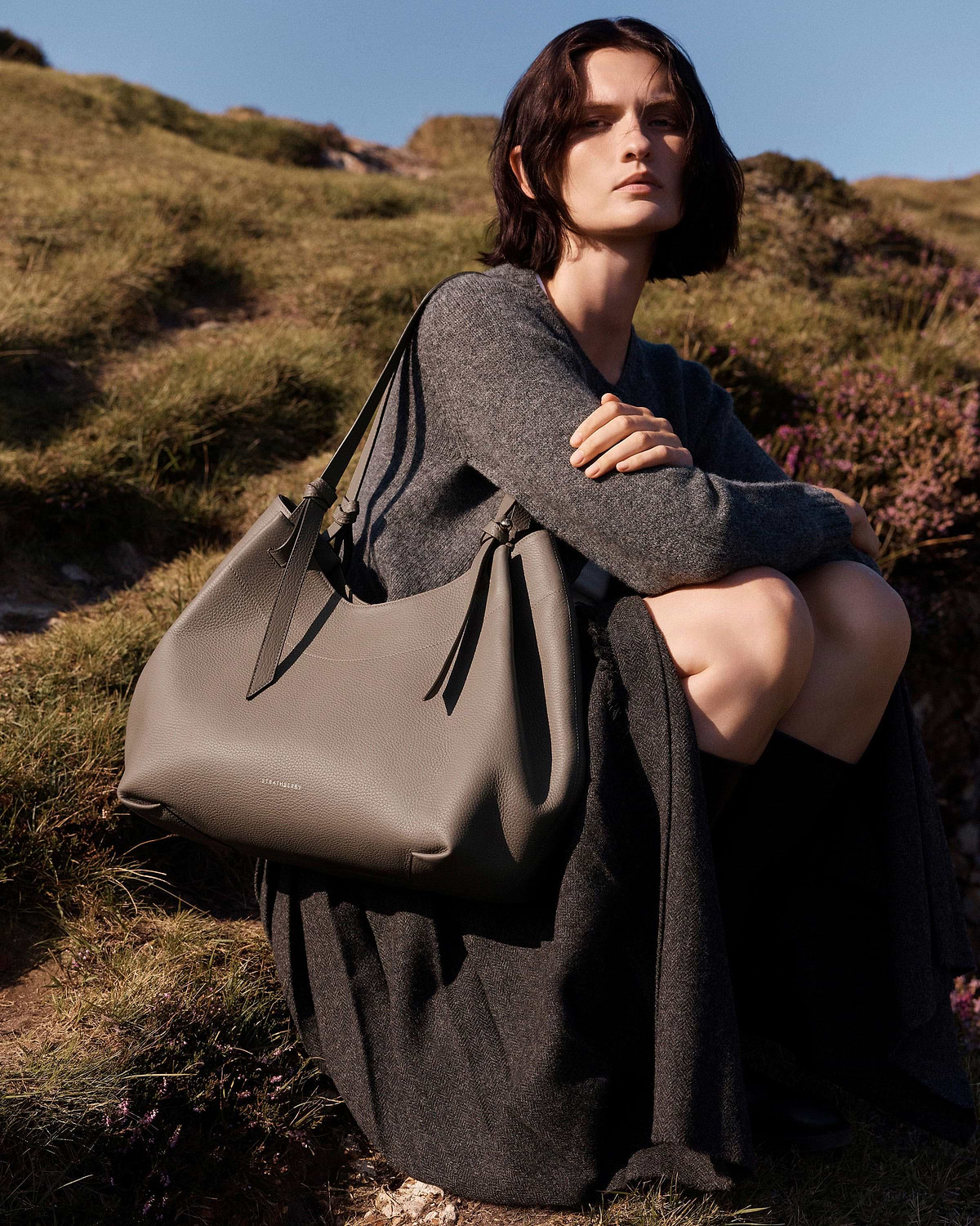 Model sitting outdoors holding taupe Strathberry Barra tote over shoulder.