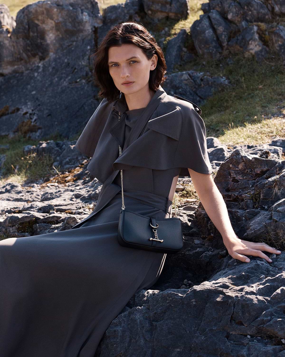 Woman in grey dress seated on rocks wearing a small black chain-strap bag.