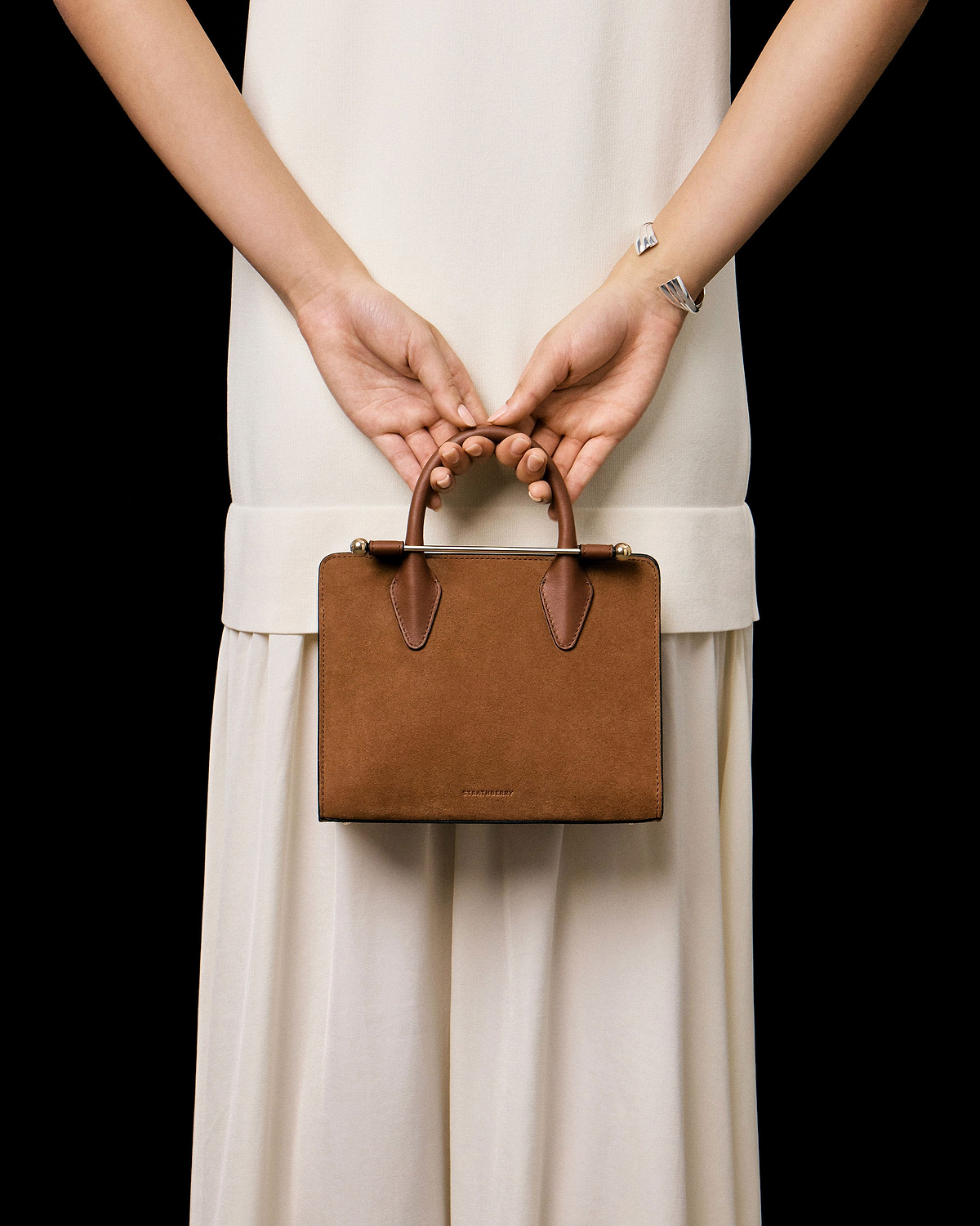 Hands holding a tan suede Strathberry mini tote against a cream dress.