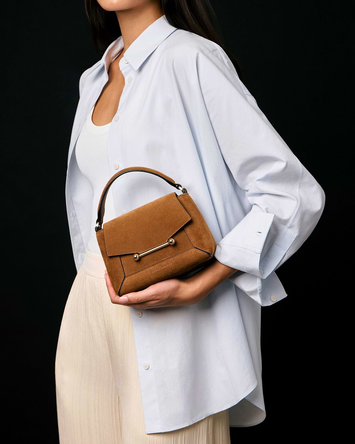 Model wearing white shirt holds tan suede Strathberry Mosaic Nano handbag.