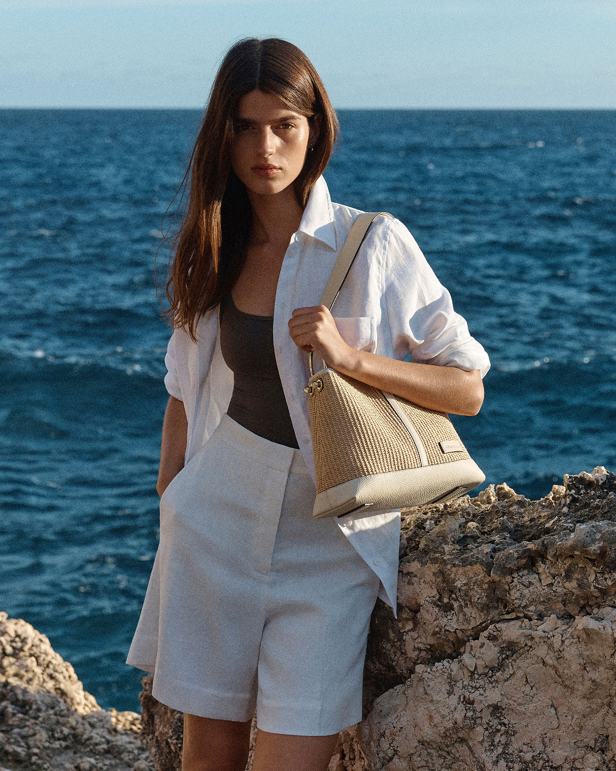 Woman in white shirt holding a vanilla natural raffia hobo bag by the sea.