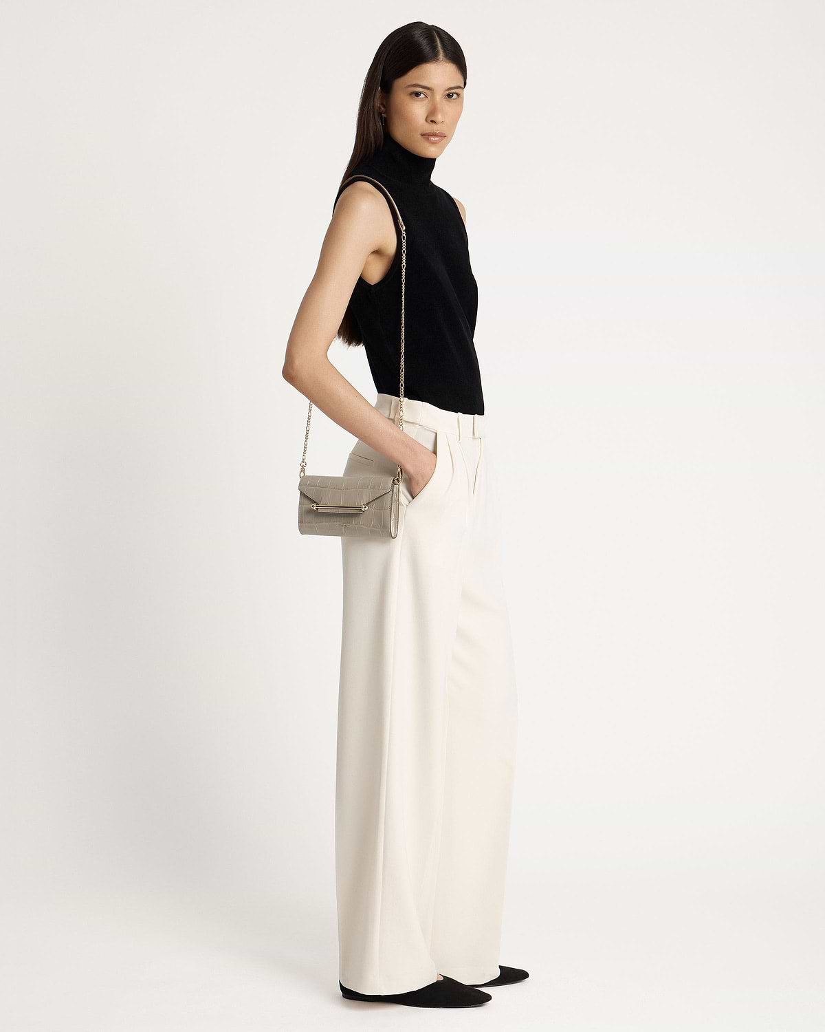 Woman wearing a black sleeveless top and cream trousers, shoulder croc chain wallet.