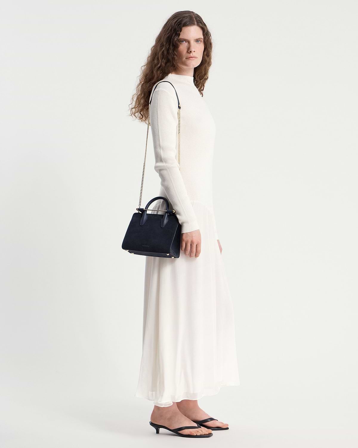 Woman in cream dress carrying navy suede mini tote with gold chain strap.