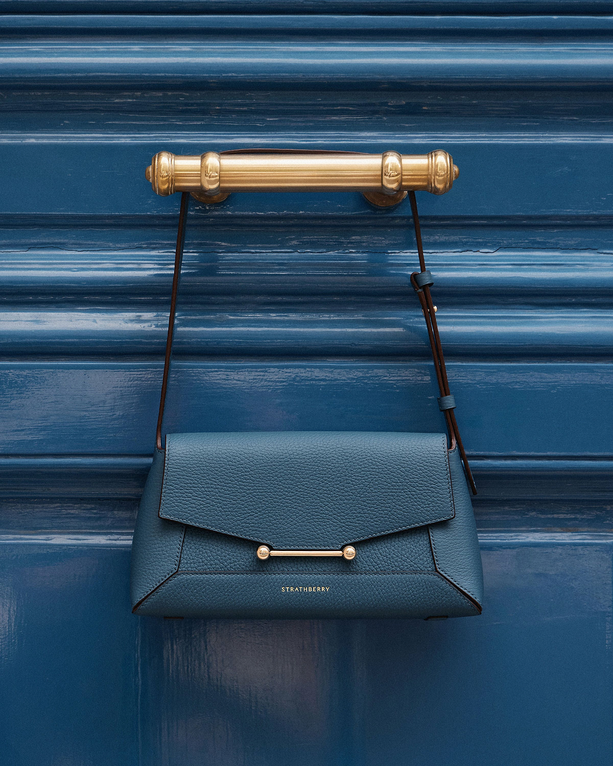 Strathberry Mosaic shoulder bag in loch blue hanging from brass door handle.