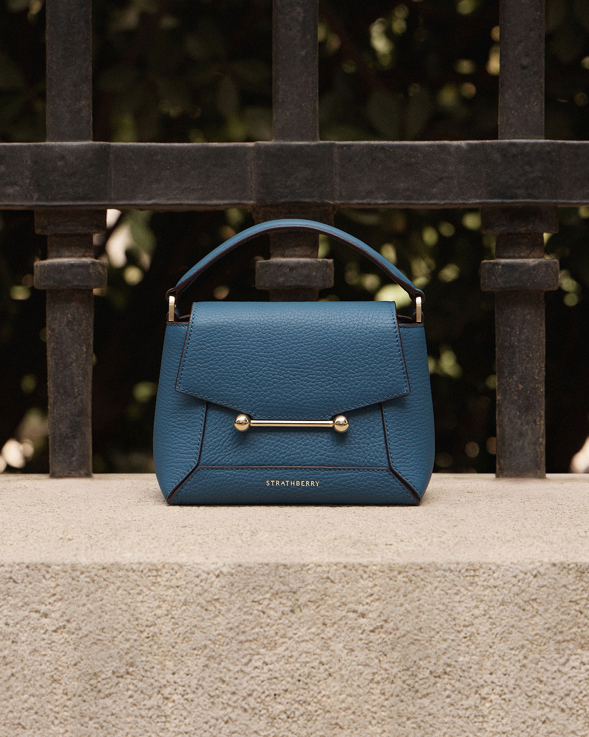 Loch blue Strathberry Mosaic Nano bag on stone ledge against iron railings.