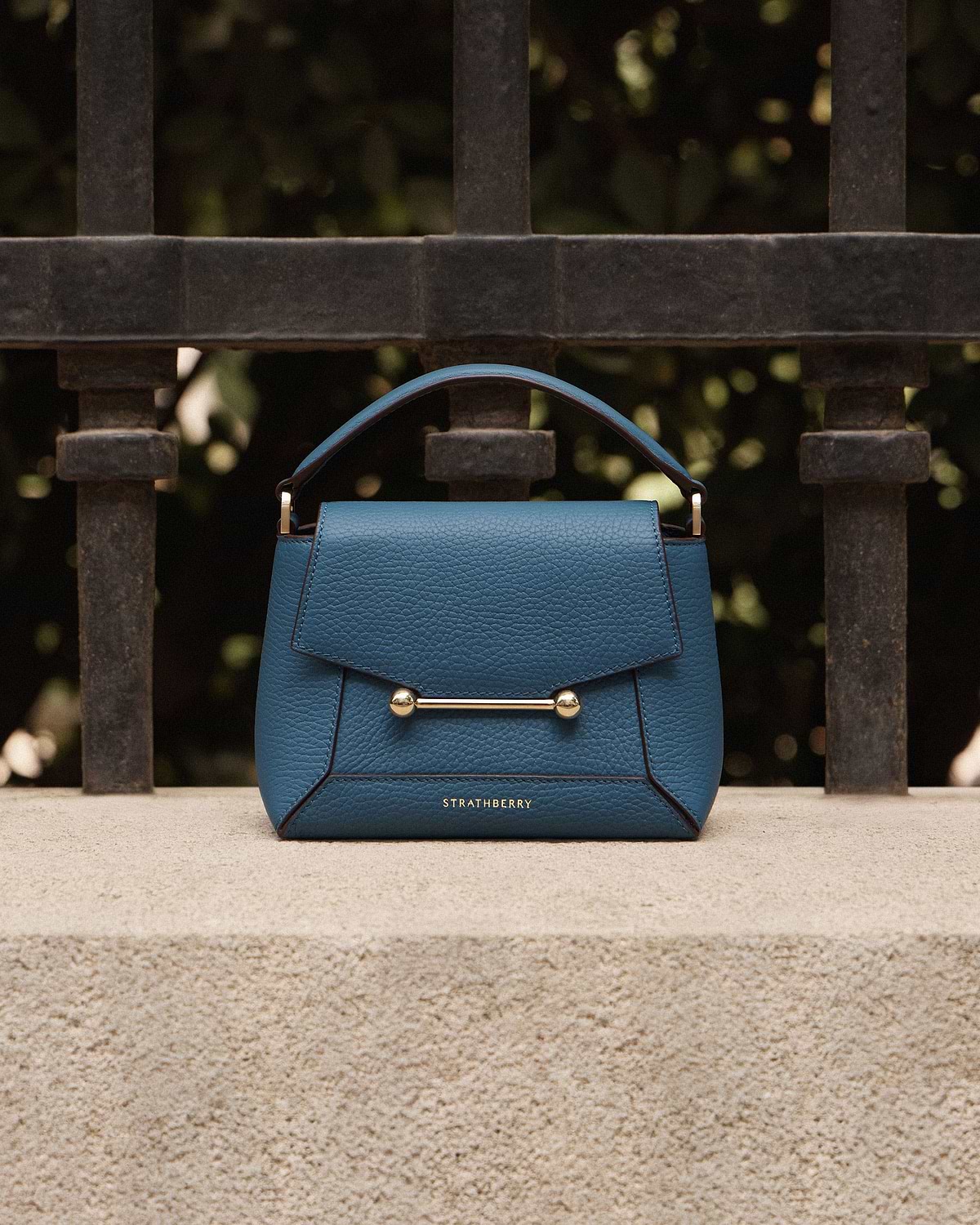 Loch blue Strathberry Mosaic Nano bag on stone ledge against iron railings.