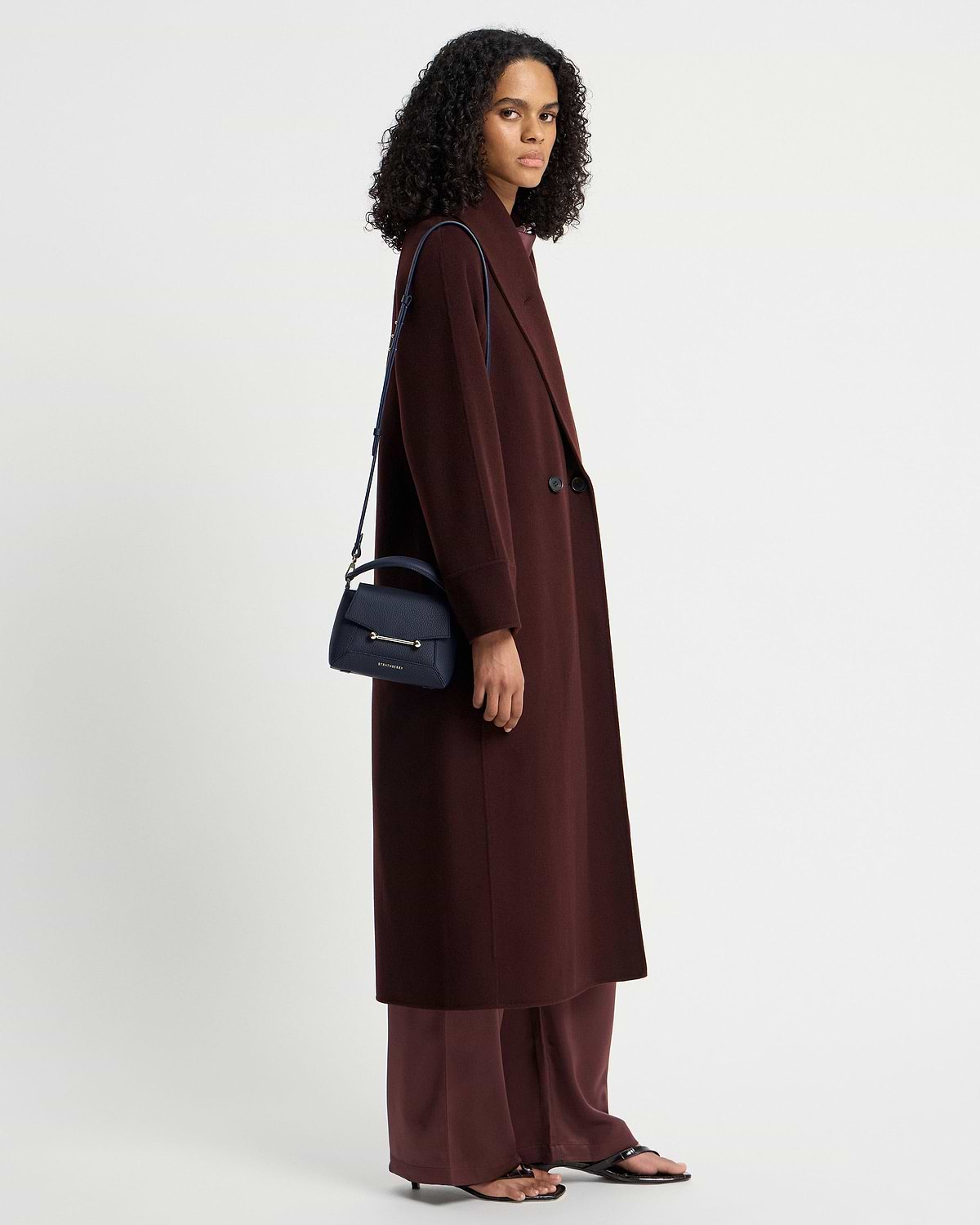 Woman in burgundy coat carrying navy Strathberry Mosaic Nano shoulder bag.