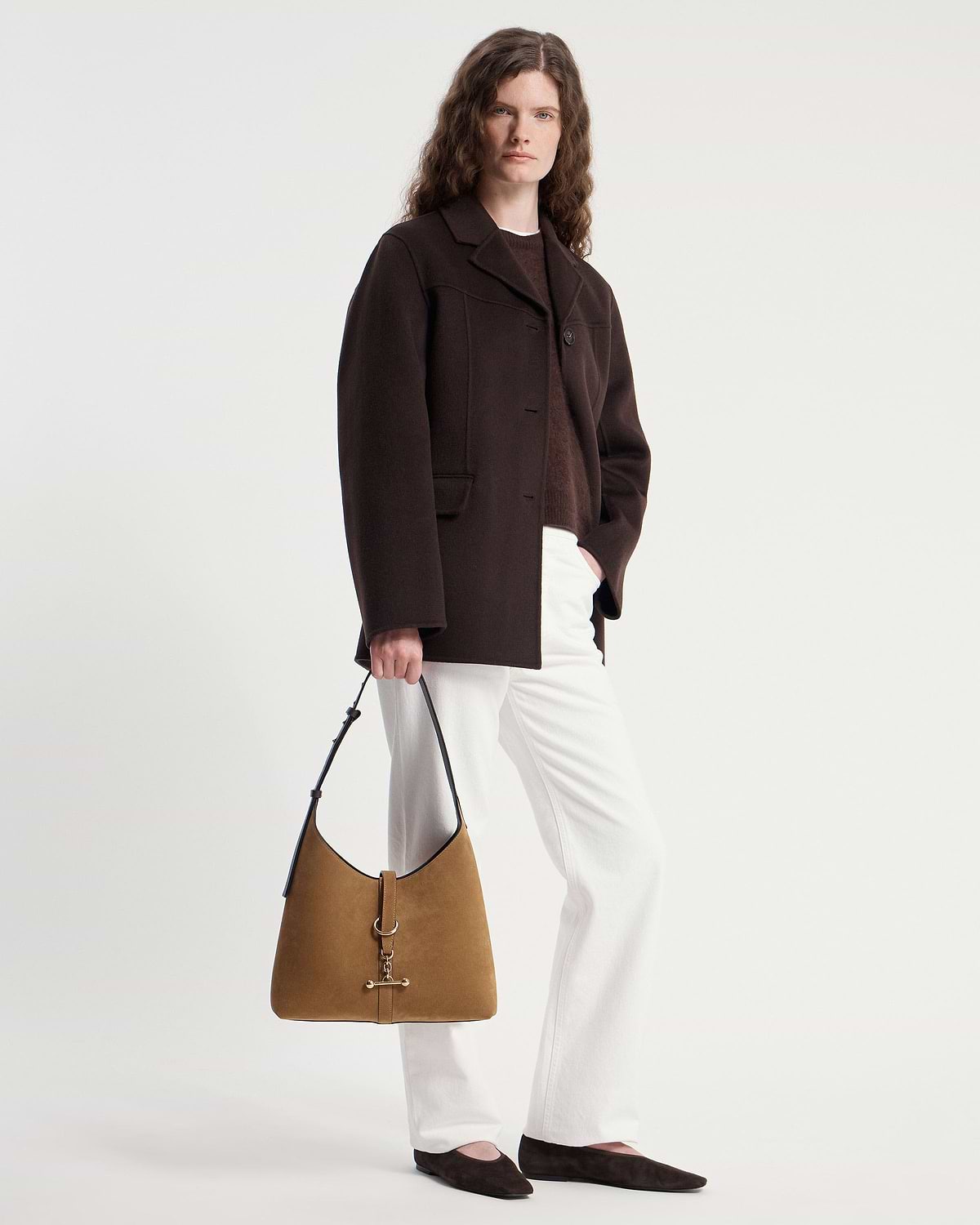 Model holding Kite Hobo toffee suede bag with espresso strap, dark coat and white trousers
