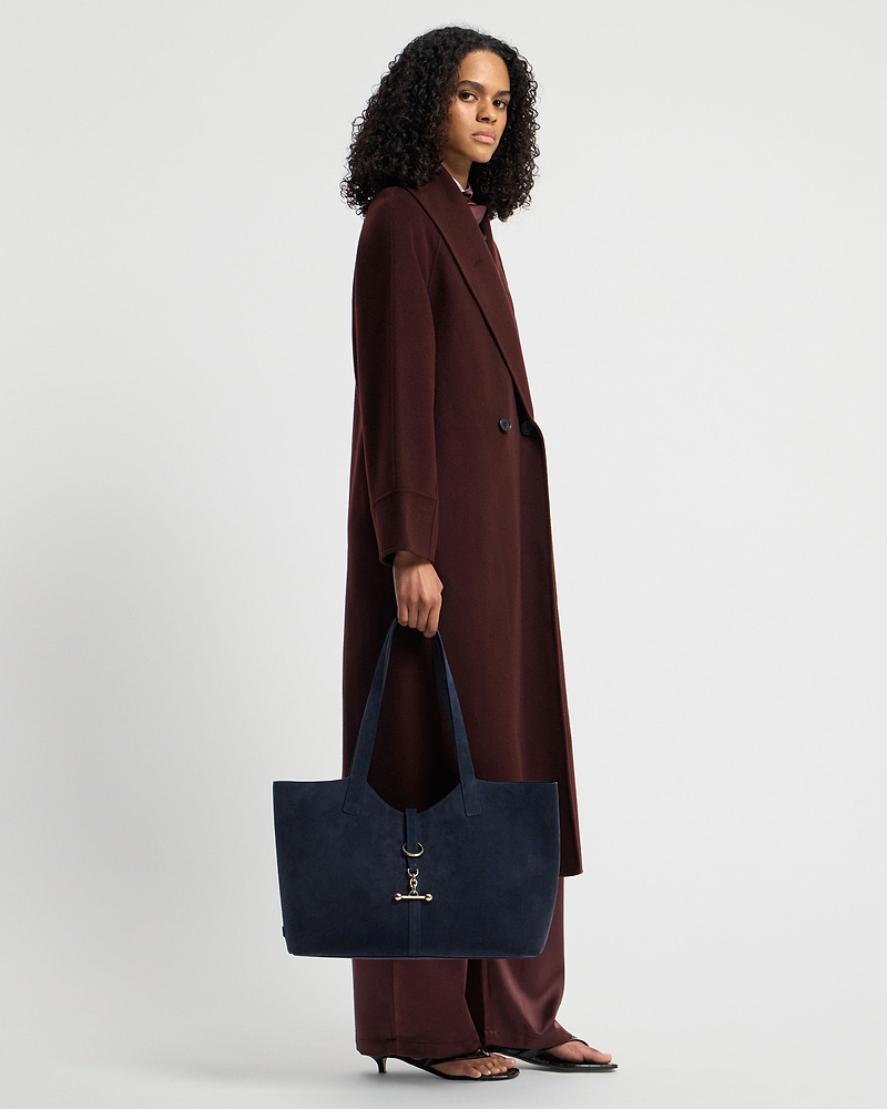 Model holding navy suede kite tote with gold hardware at her side, wearing long coat.
