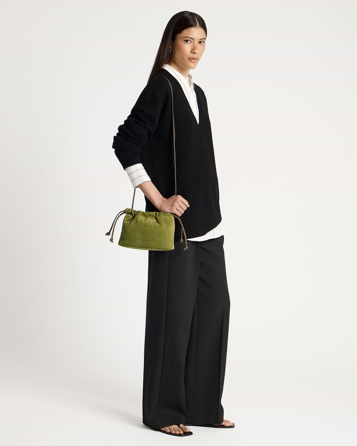 Model wearing black V-neck sweater holds lime suede drawstring bag at her hip.