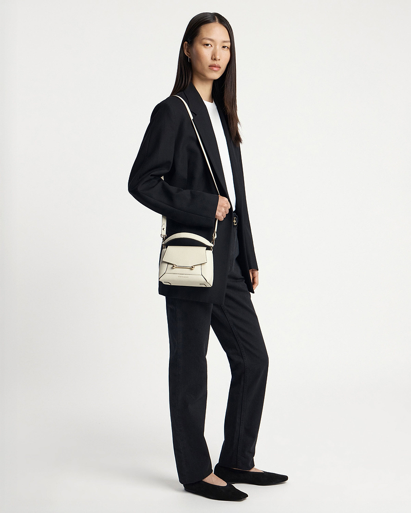 Model wearing a small vanilla crossbody bag with top handle and metal bar.