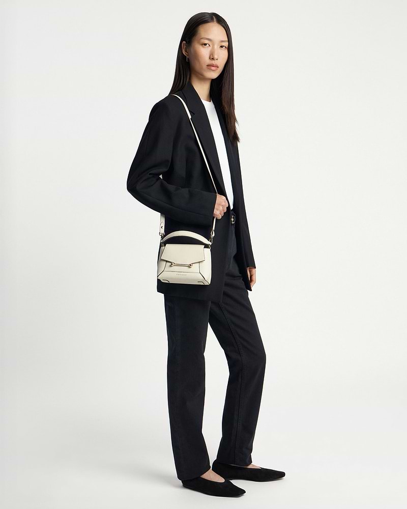 Model wearing a small vanilla crossbody bag with top handle and metal bar.