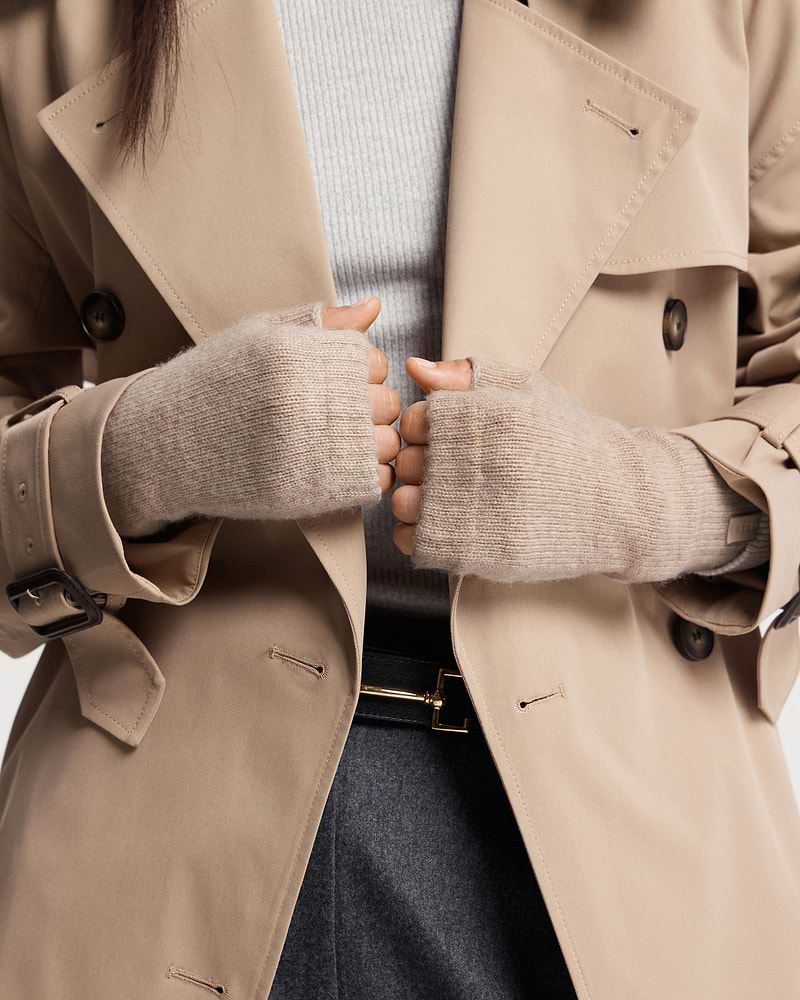 Hands wearing oat fingerless cashmere gloves holding beige trench coat lapels.
