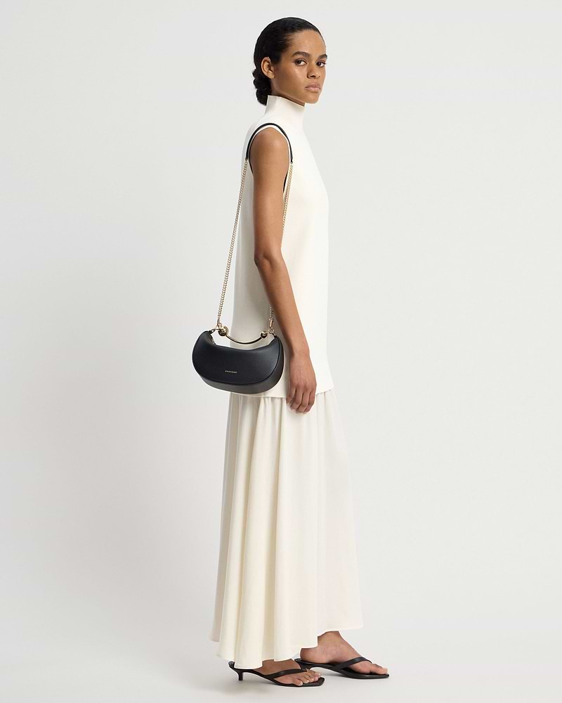 Model in white sleeveless dress carrying black Strathberry Crescent Moon Mini shoulder bag