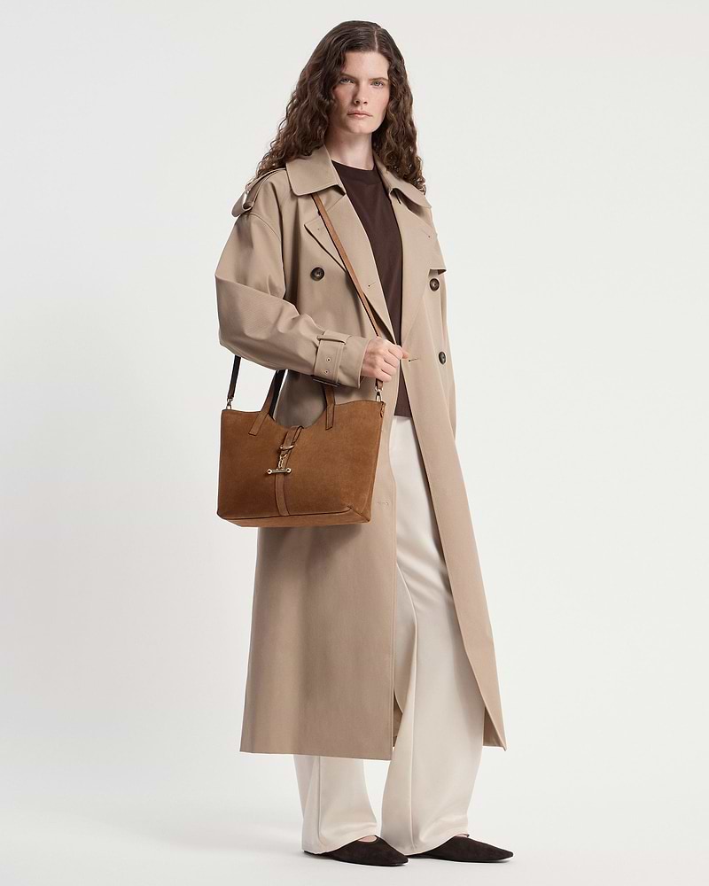 Model in a beige trench coat carrying a tan suede tote bag at their side.