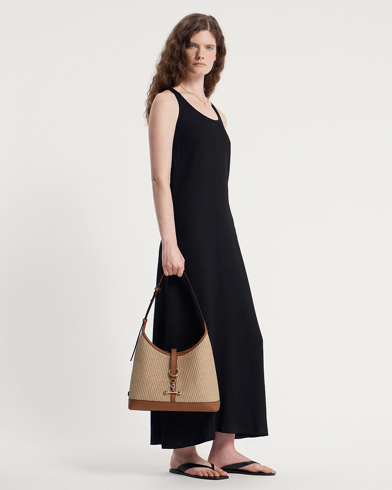 Model wearing black sleeveless dress holding tan raffia hobo bag with leather trim.