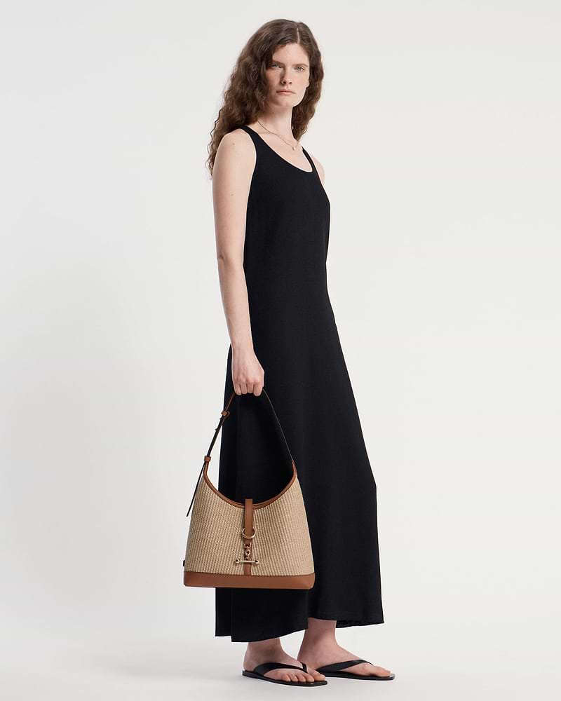 Model wearing black sleeveless dress holding tan raffia hobo bag with leather trim.