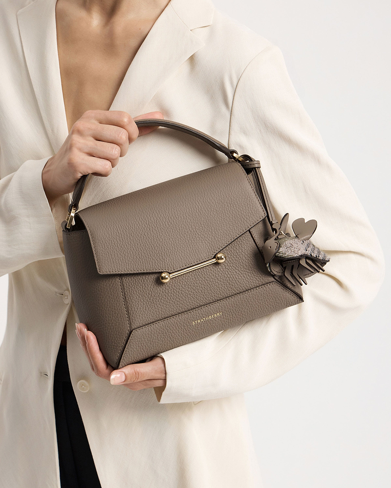 Model holding taupe Strathberry leather bag with tartan bumblebee bag charm.