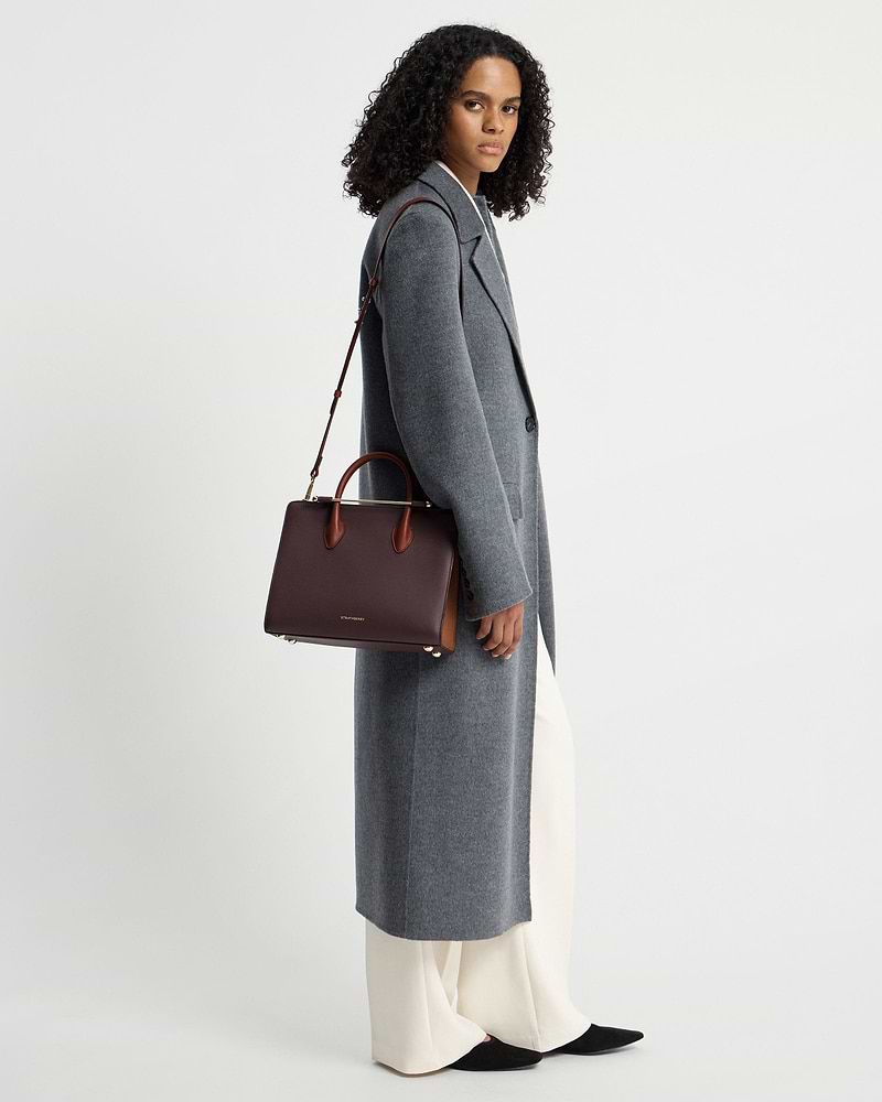Model wearing grey coat holding Strathberry midi tote in burgundy, walnut, chestnut.