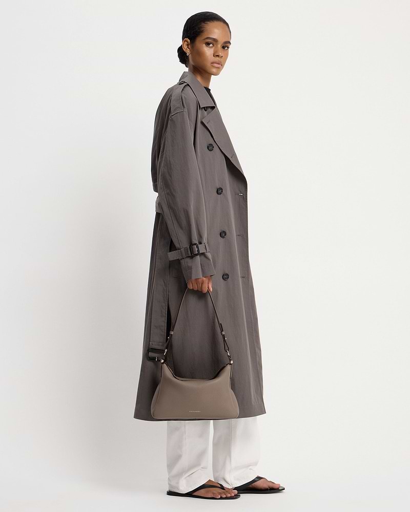 Model wearing taupe Strathberry Sway Maxi bag with trench coat and white trousers.