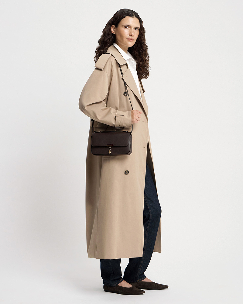 Model in trench coat wearing espresso Nocturn clutch with gold charm and shoulder strap.