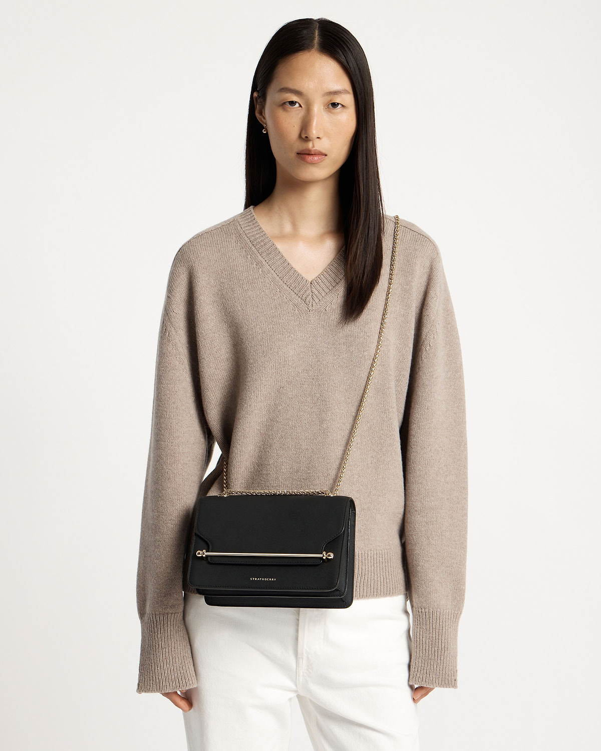 Model wearing black Strathberry east-west crossbody bag with gold bar and chain strap.