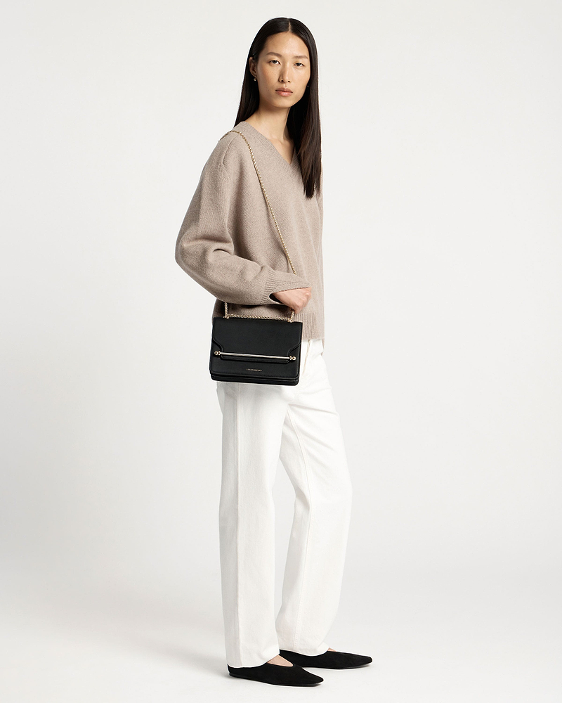 Model wearing black east-west shoulder bag with gold chain and boxy flap.