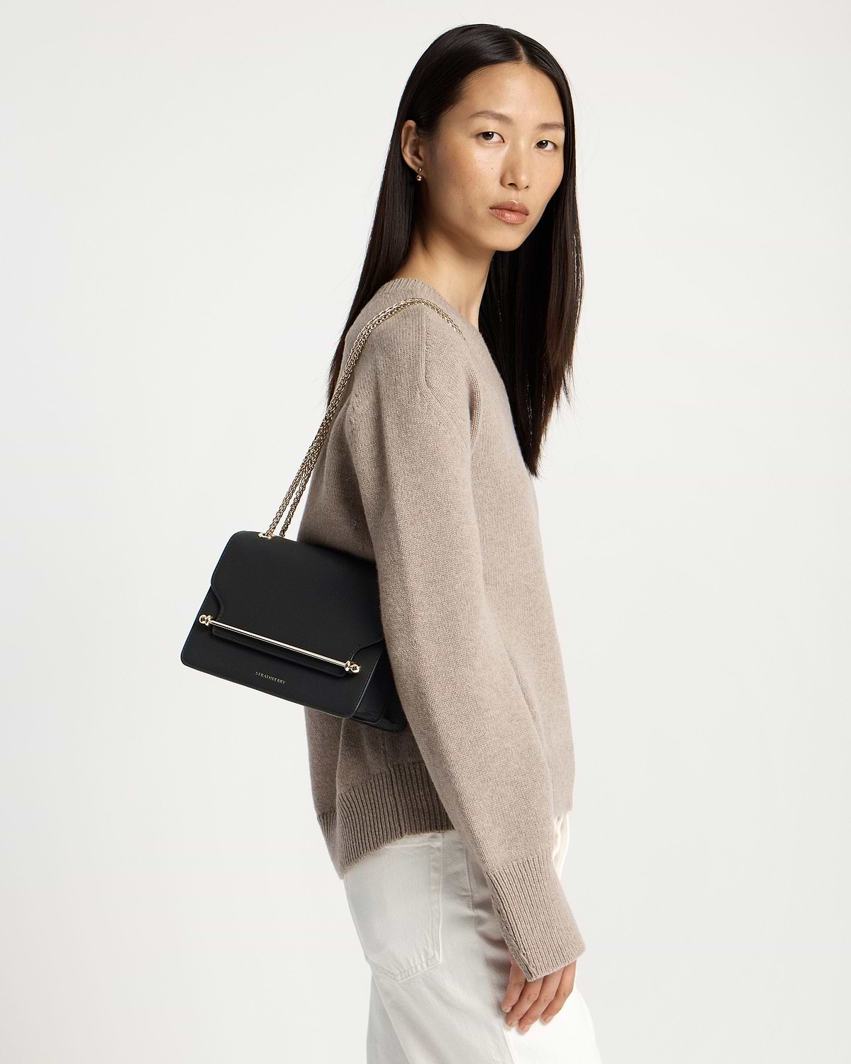 Model wearing beige sweater carrying black east-west bag with gold chain strap and bar.