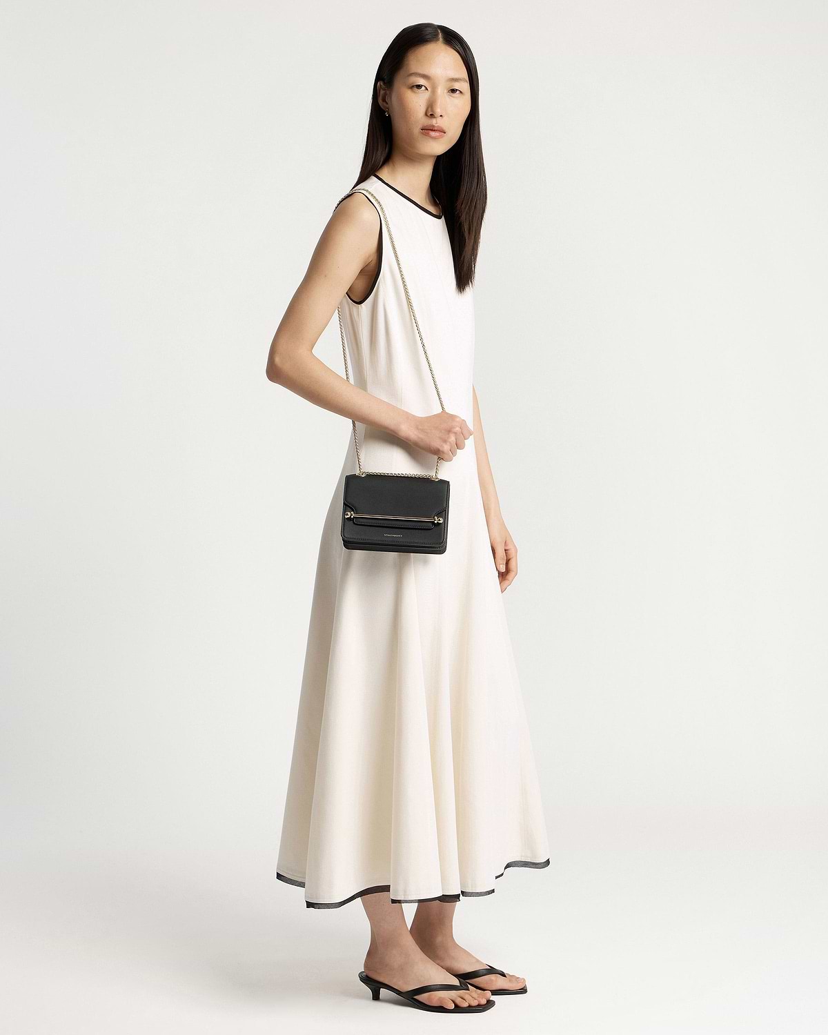 Model in a white sleeveless dress holding a small black crossbody bag with chain.