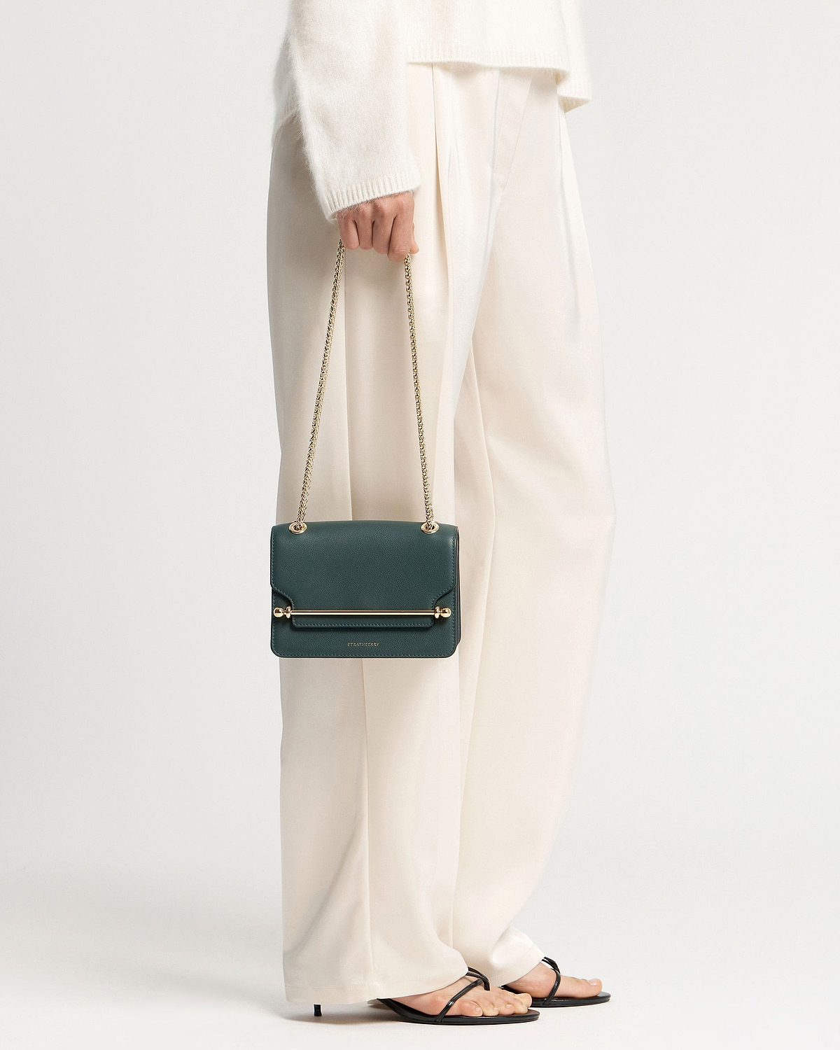 Model holding bottle green Strathberry East/West Mini bag with gold chain strap.