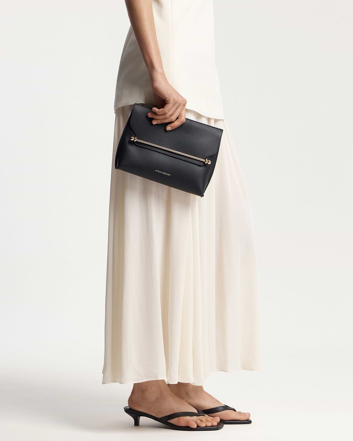 Model waist down holding black Strathberry Stylist clutch with gold bar.