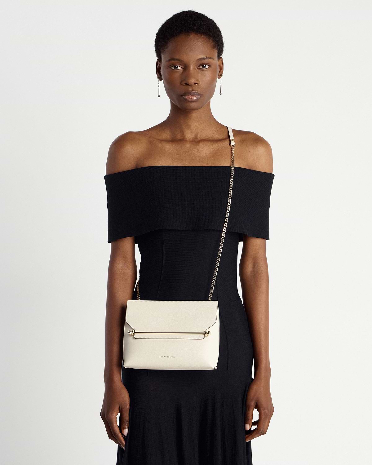 Model in black off-shoulder dress wearing Strathberry Stylist bag in vanilla with gold chain.