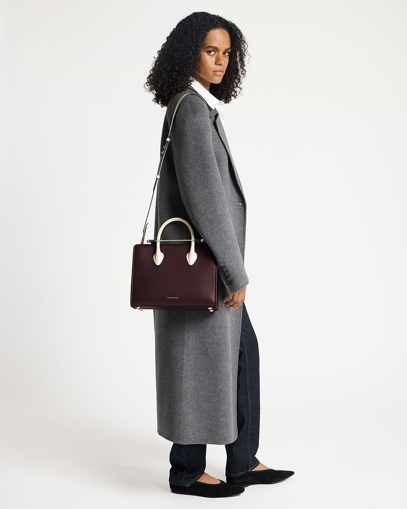 Model wearing a grey coat holding a Strathberry midi tote in burgundy, navy and vanilla