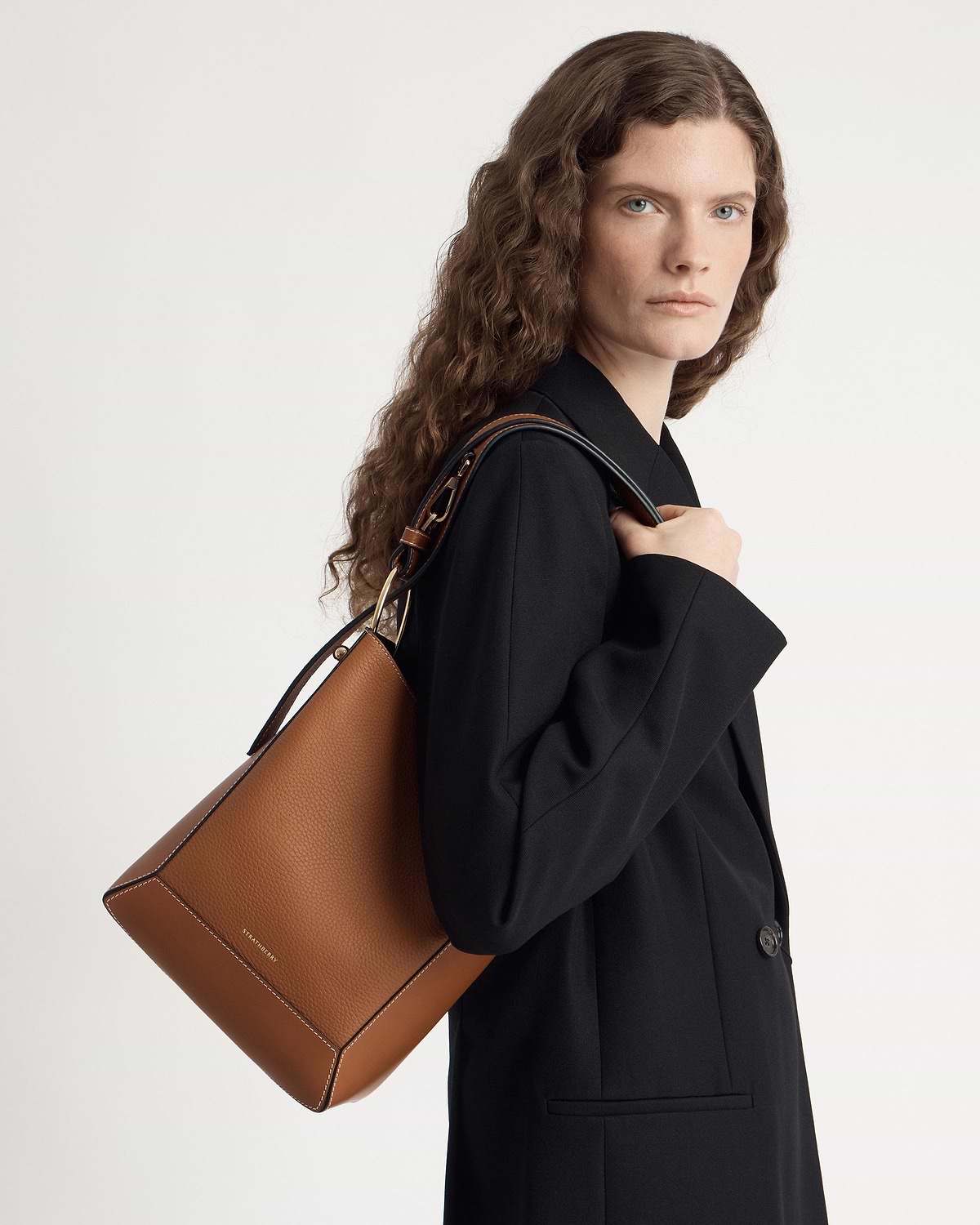 Model in black blazer carrying Strathberry midi bucket bag in tan with vanilla stitch.