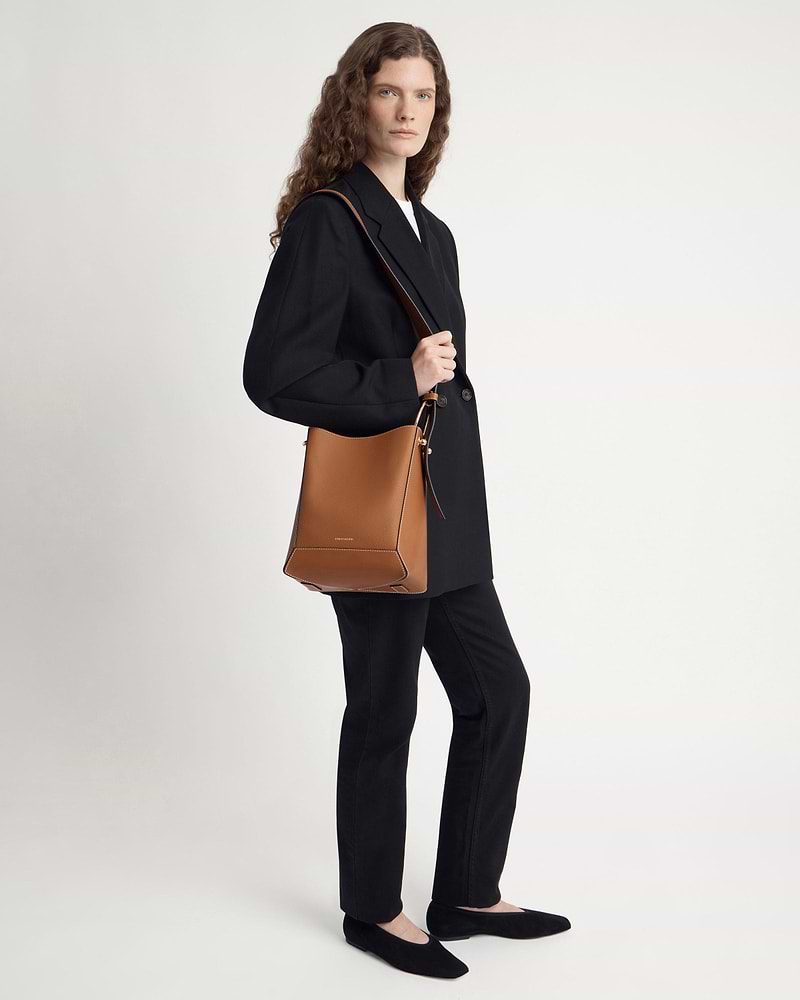 Model standing wearing black blazer and trousers, carrying tan bucket bag with vanilla stitch.