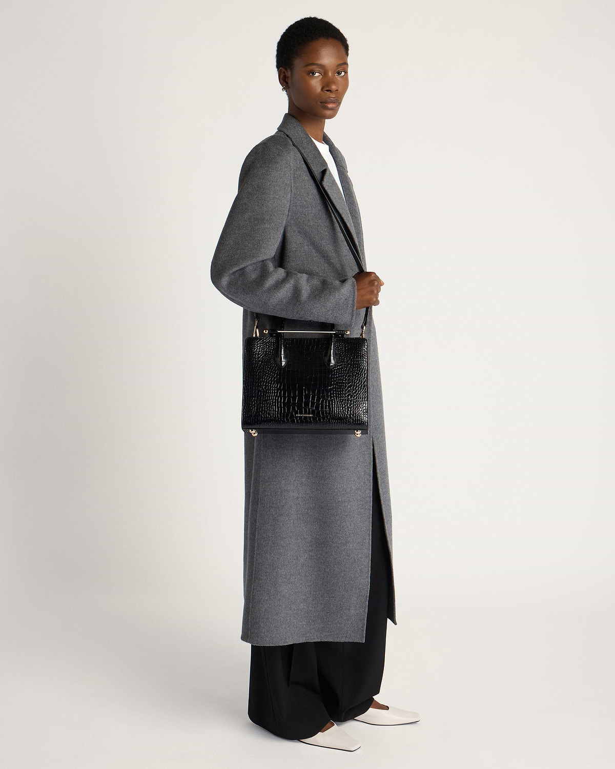 Model wearing grey long coat holding black croc midi tote bag at side.