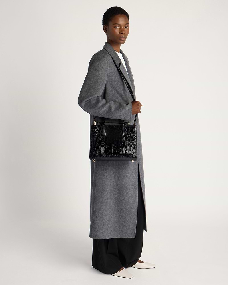 Model wearing grey long coat holding black croc midi tote bag at side.