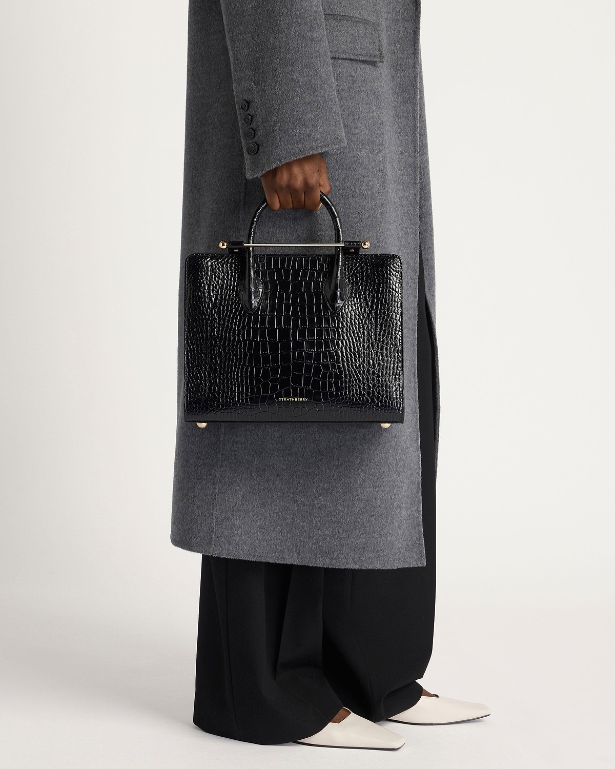 Model holding Strathberry midi tote in black crocodile-embossed leather, worn with grey coat.
