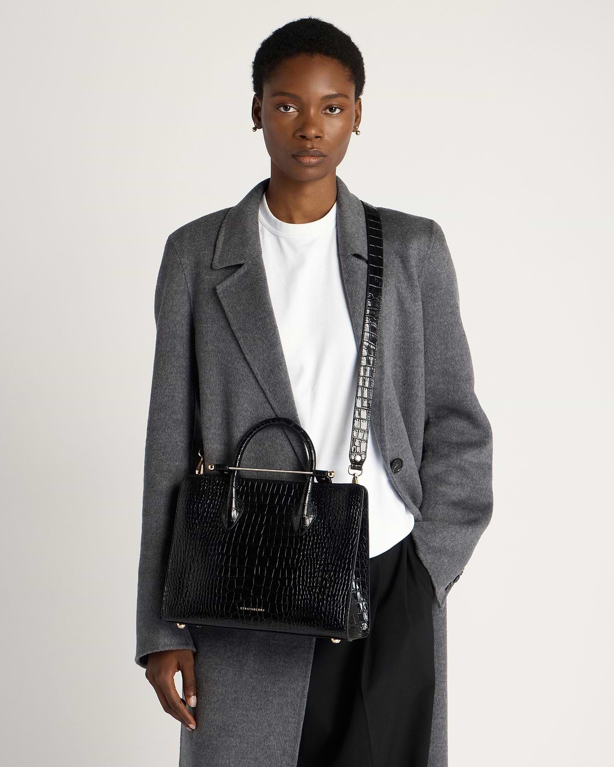 Model wearing grey coat carrying Strathberry black croc midi tote with shoulder strap.