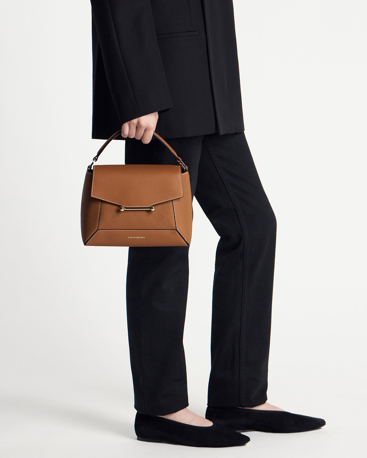 Model cropped below waist holding tan Mosaic bag with vanilla stitching, Strathberry logo visible.