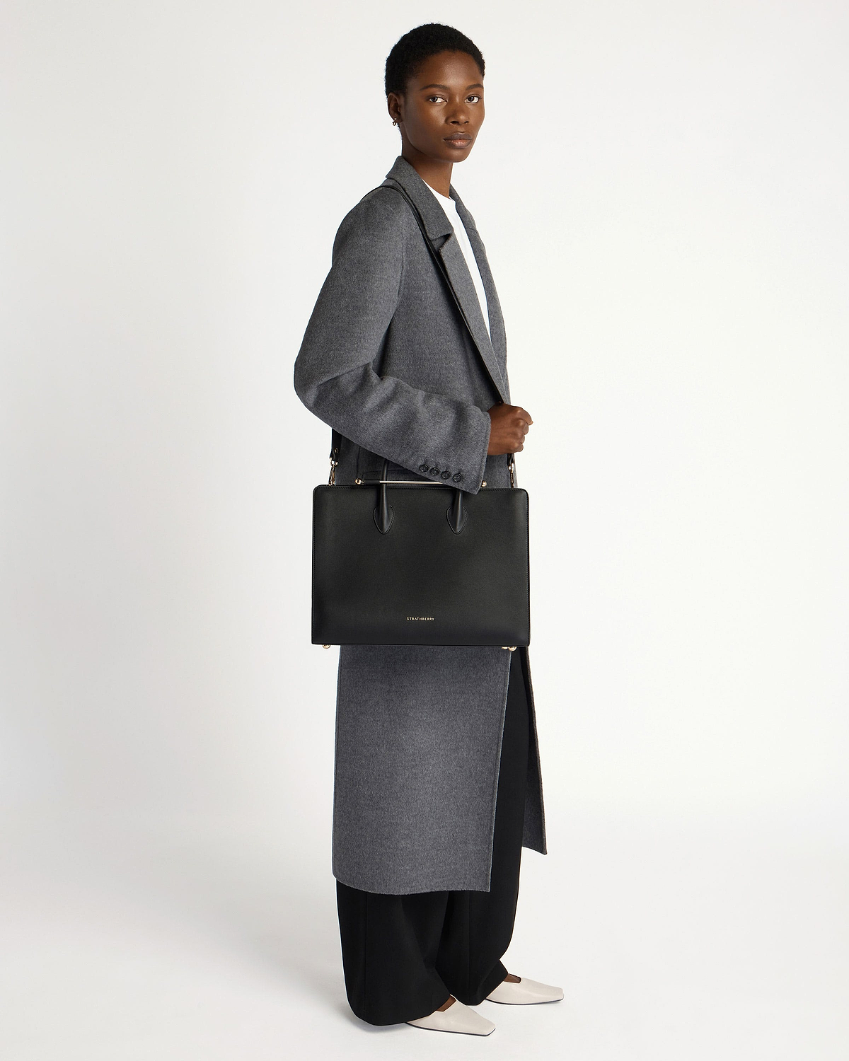Model wearing grey coat holding black Strathberry tote bag over shoulder.